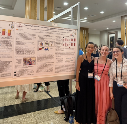 Three people stand beside a research poster at an academic conference, smiling at the camera. Wearing conference name badges, they pose in a busy poster session with other presenters and attendees in the background.